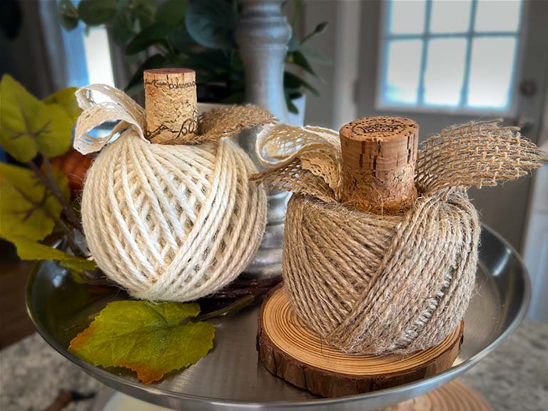 Two balls of twine are made into pumpkins by adding white ribbon and burlap cut into leaves with a cork for a stem.