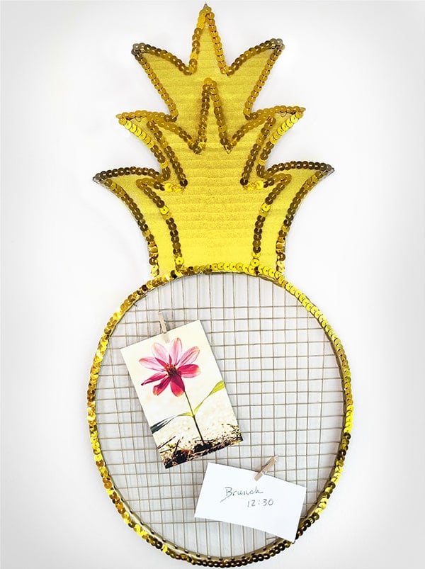 Vertical photo of a DIY pineapple memo board made out of a dollar store badminton racket, painted gold with a cardboard top and gold sequins around the edges.