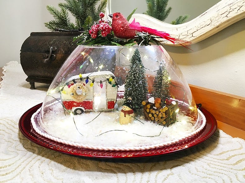 Photo of a finished snow globe scene made from a large plastic bowl and red charger plate. Features a mini camper, fairy lights, bottle brush trees a snowman and wood pile.