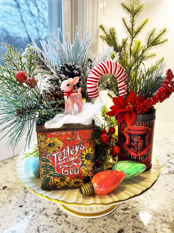 Tray filled with greenery, vintage colorful Christmas light bulbs, a Rumford Baking Soda can with greenery in it and a rectangular vintage Tetley's Tea container with greenery, faux snow and a small pink vintage looking reindeer in it.
