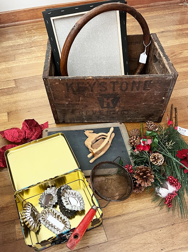 Shown is an old wood crate and thrift store items I collected to upcycle into Christmas decor items to sell in my antique booth! There are florals, an old red handle strainer, a tin box, tart tins, and a vintage chalk board.