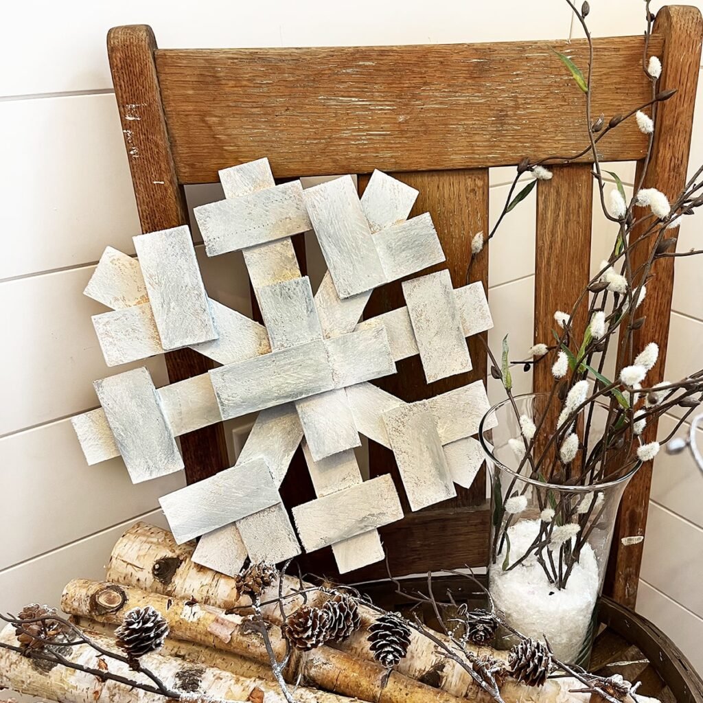 Square feature of a wood snowflake decoration sitting on a chair on top of a basket of birch logs and pinecones with a clear glass vase of willows on the side.