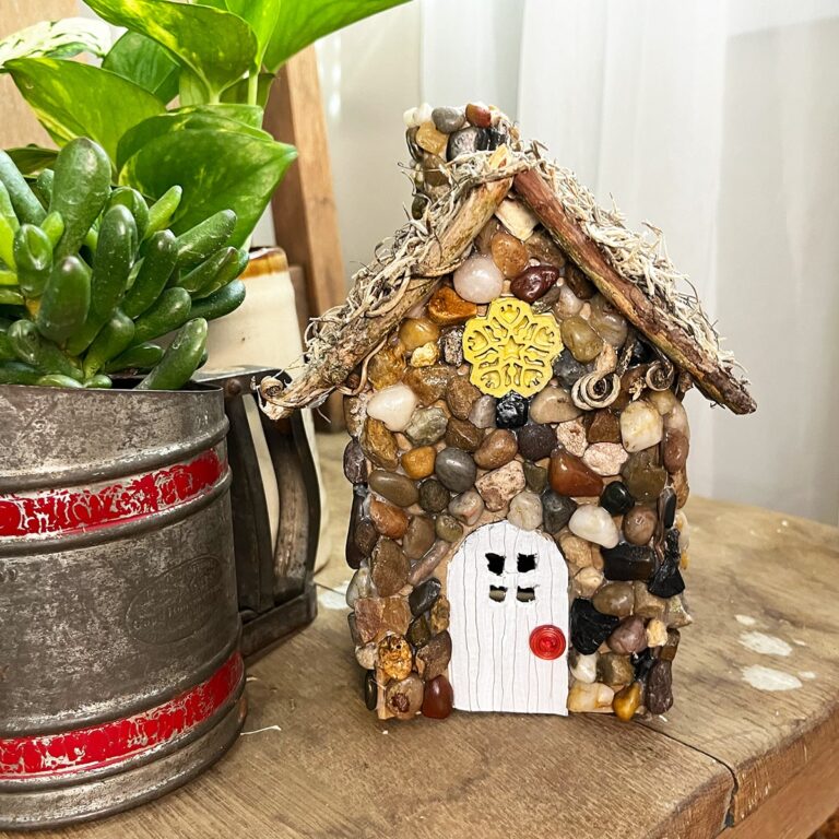 Square feature photo of a finished stone pebble fairy garden house on a wood chair with a plant in a vintage sifter on the left side.