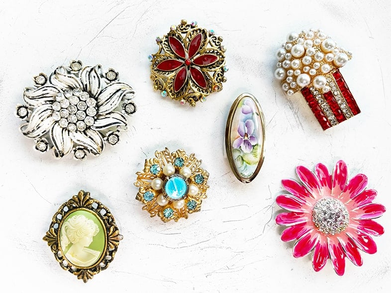 Photo of seven colorful brooch pins, some new and some vintage. There is a popcorn pin, floral pins, and a cameo pin.