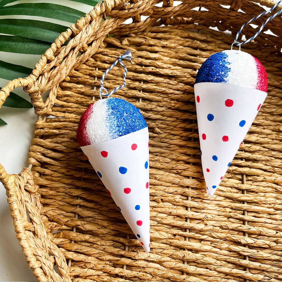 Finished DIY snow cones in red, white and blue for the snow top and red and blue dots on a white snowcone cup. String hanging from the top but they are shown in a wicker basket with a palm leaf behind for Christmas and July!