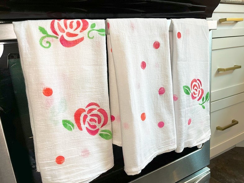 Photo of three finished hand-painted tea towels with pink shades of roses hanging on a oven range handle.