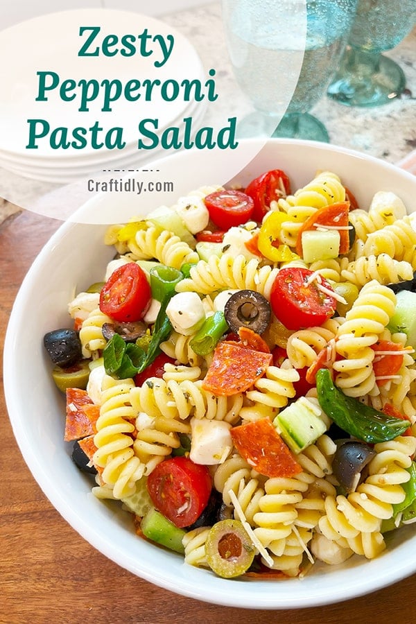 A vibrant photo of a white serving bowl with zesty pepperoni pasta salad. You can see green onion, fresh basil leaves, pepperoni, vibrant banana peppers, black and green olives and grape tomato halves all glistening in Italian seasonings and dressing. This is a vertical image to pin to your Pinterest recipe boards to come back to this summer favorite recipe later!