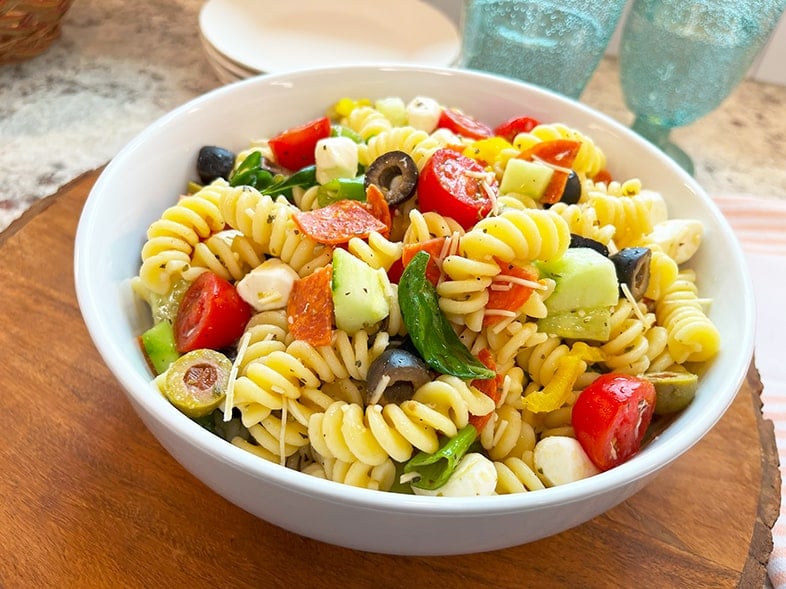 A vibrant photo of a white serving bowl with zesty pepperoni pasta salad. You can see green onion, fresh basil leaves, pepperoni, vibrant banana peppers, black and green olives and grape tomato halves all glistening in Italian seasonings and dressing. Bright and colorful with summer sparkly aqua glasses in the background.