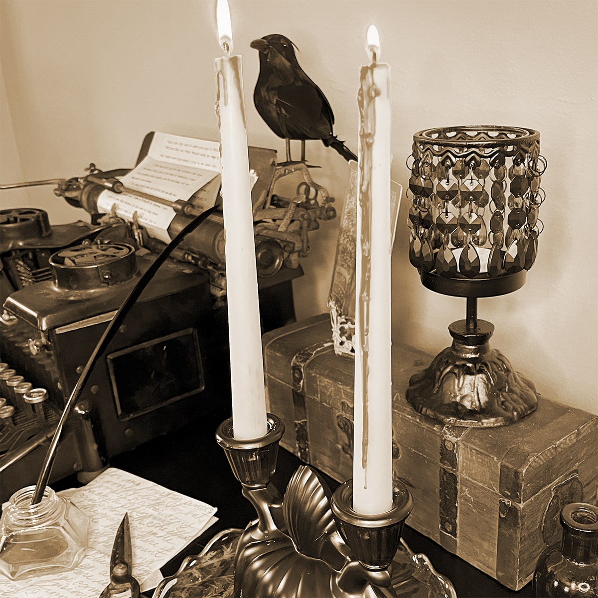 Square feature photo of a vintage Halloween vignette with a crow on a vintage typewriter and candleabra with fake wax blood dripping down in the foreground on a black desk. There is a handwritten letter to the side with vintage scissors and a jar with an old quill black feather pen. The tone is spooky and eerie and the photo is sepia.