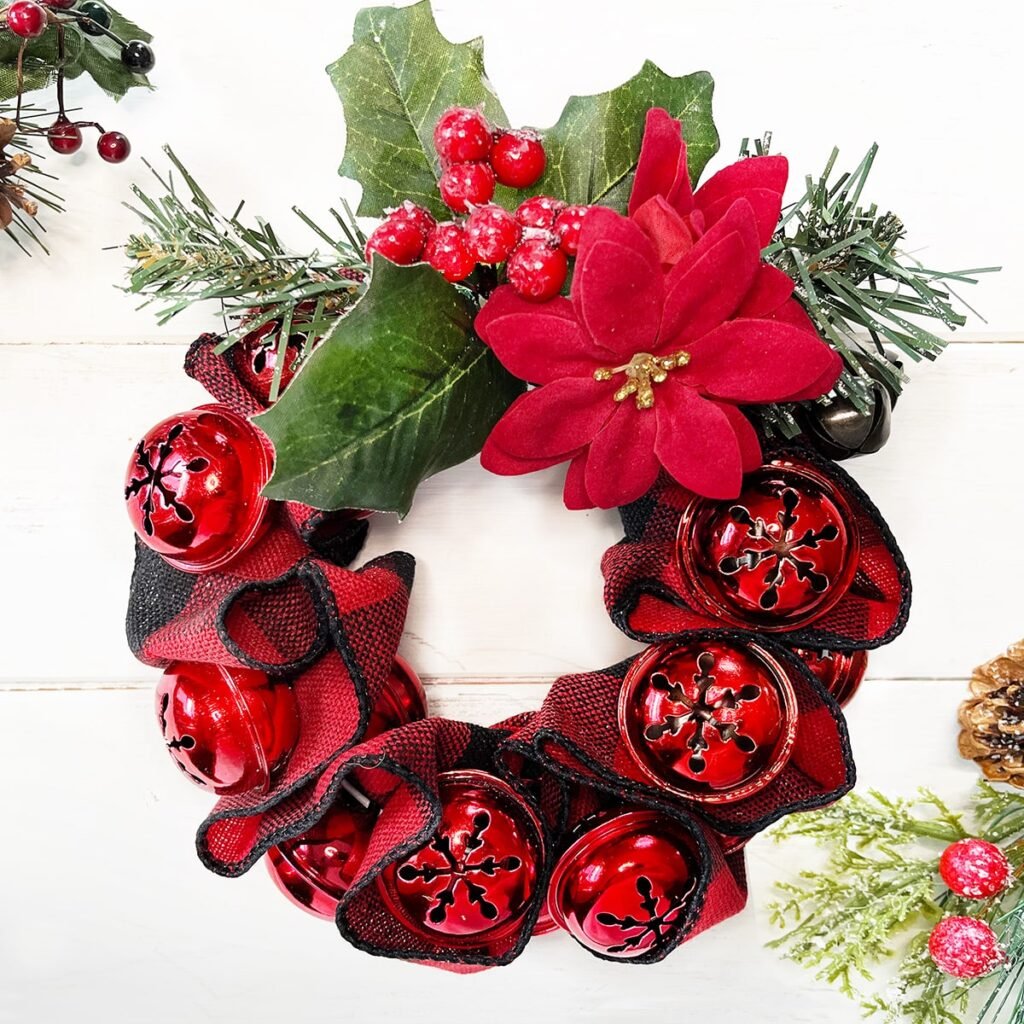 Square feature photo of a finished Jingle Bell & Ribbon wreath on a white wood background. This is a small sized wreath made with large decorative bells, 2-inch red and black check ribbon and a wire hanger. The decoration at the top is a red berries and red velvet Poinsettia pick.