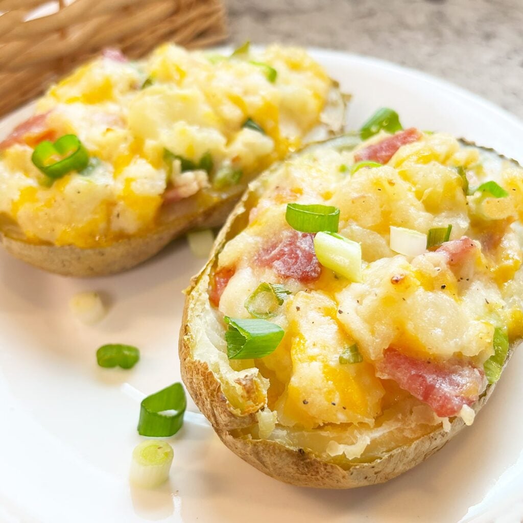 Photo of two stuffed twice-baked potatoes with bacon hot from the oven on a white plate. In background is another plate with more and a wicker basket. The potatoes have cheddar cheese mixed into the potato with green onions. bacon bits and seasoning mixed in. All are heaped full in the potato skin boats. Slightly brown on top but soft and delicious on the inside.
