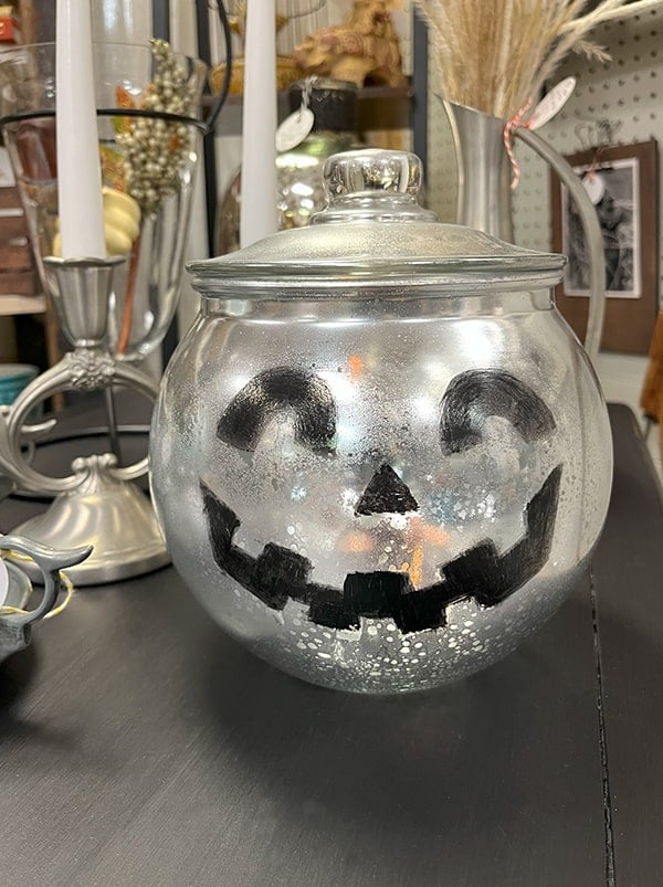 Jack o lantern made from an thrift store round clear glass cookie jar with a lid. The jar was spray painted to look like mercury glass in silver and a smiling Jack-O-Lantern face painted on the front with black enamel paint. The glass jar has a tea light inside and is sitting on a black table in my antique booth to greet customers.