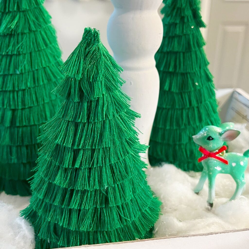 Closeup square feature photo of three mini Christmas Tree decorations made with fringe ribbon and a cardboard base. Shown on a snow drape in white with a tiny green deer with a red bow around its neck to the side.