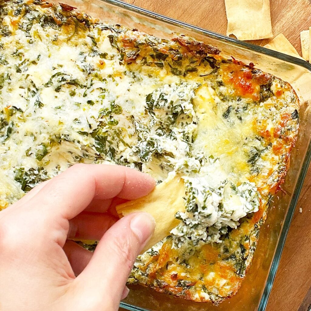 Photo of creamy spinach dip served in a clear baking dish on a wood board with pita chips in the background. This dip is served warm and is golden brown around the edges and topped with gooey cheese and green spinach throughout. Here there is a closeup of a hand dipping a pita chip into the dip just out of the oven.