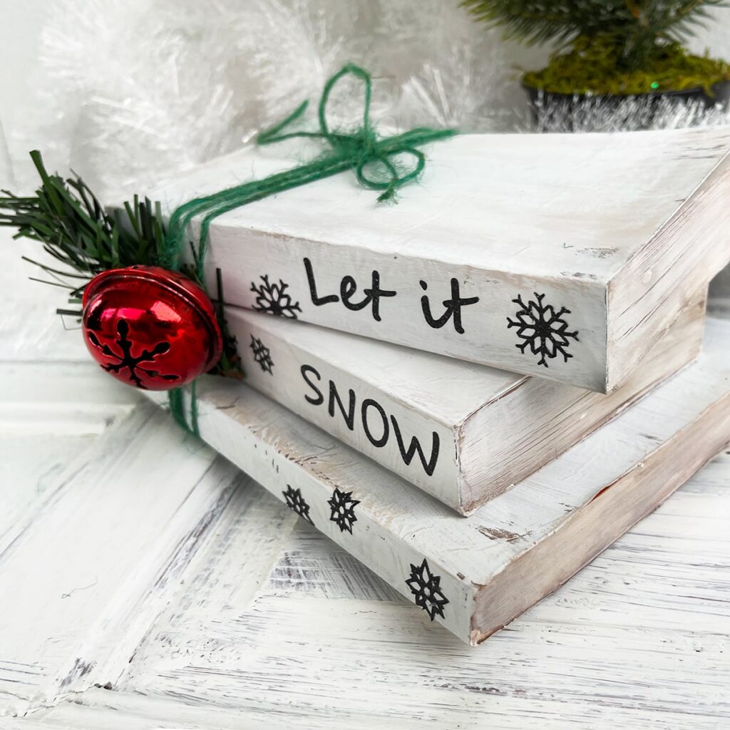 Photo of a white chalk painted book stack with 'Let it Snow' on the binding and snowflakes art in black vinyl cut on a Cricut Maker. The three books are tied together with green jute and there is a big red bell and a sprig of pine greenery to the side. The book stack is on a white wood table with greenery and fluffy white garland in the background.