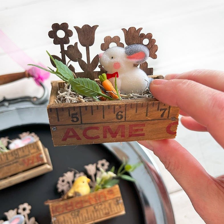 Closeup photo of a ruler box magnet made from a vintage yardstick with ACME advertising on the front and filled with floral moss, a sprig of greenery, a side profile of a white flocked bunny with gray ears, a pink nose and cheeks. In the background is a mini wood fence with cut out flowers. Next to the bunny is a mini orange carrot.