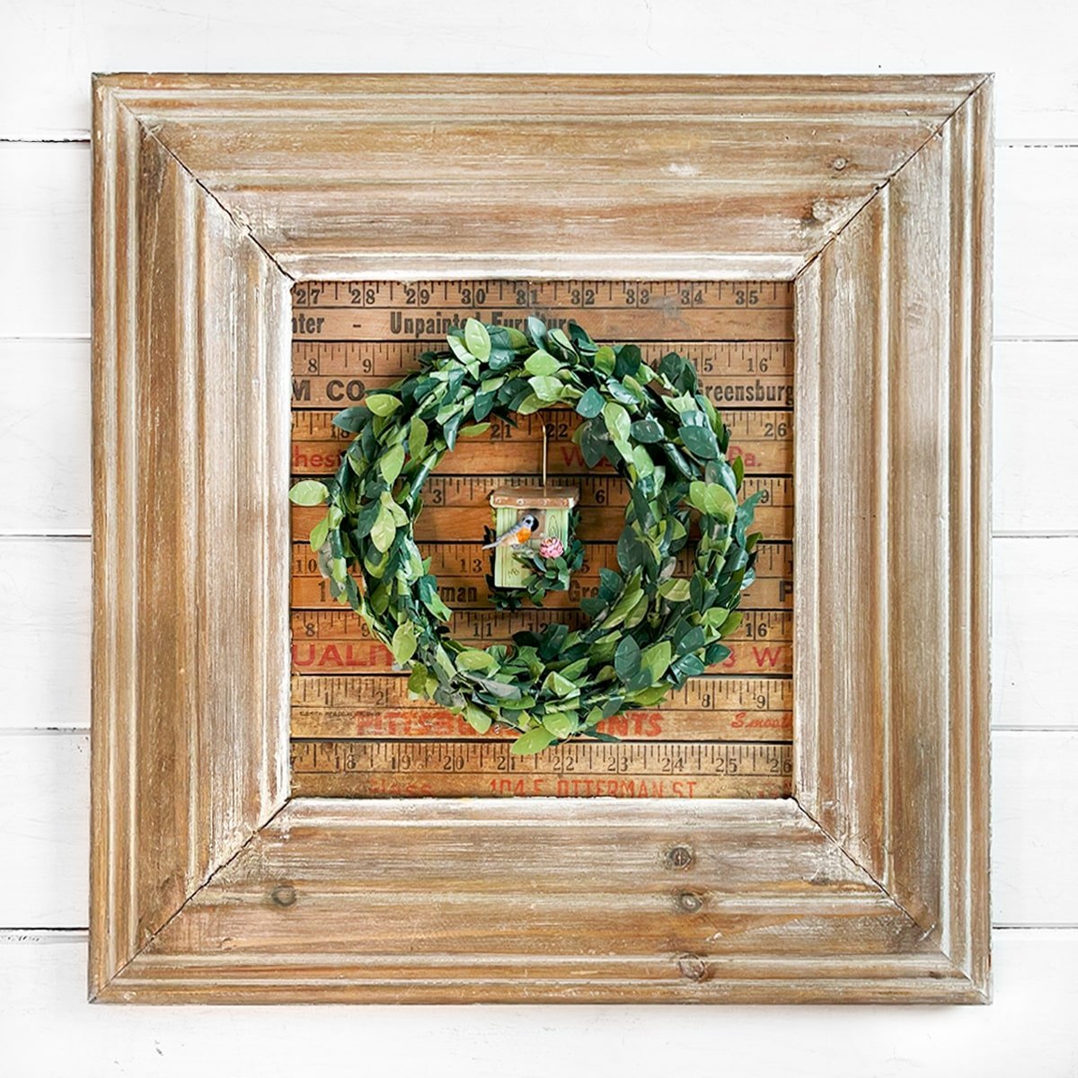 Photo of a square wide wood frame lined with vintage yardsticks. This rustic frame frame had a boxwood greenery wreath in the center and in the center of the wreath hangs a small porcelain light spring green birdhouse ornament.