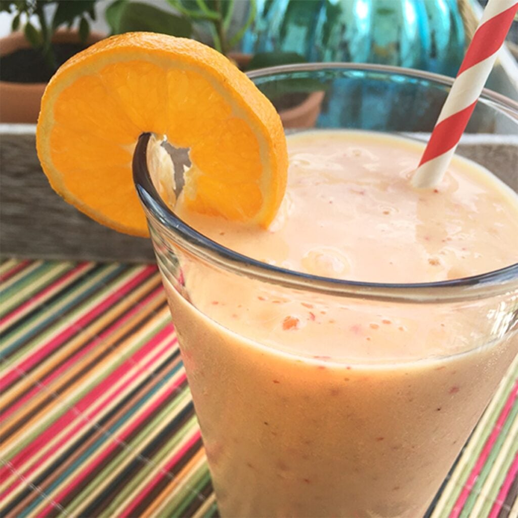 Square feature photo of a banana pineapple mango smoothy. The color is a light pinky peach. It's served in a clear glass with an orange slice on the side of the glass with a a colorful red and white straw sitting on a multicolor bamboo placement in a summery setting.
