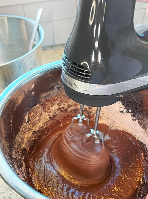 Closeup process photo of a gray KitchenAid Hand Mixer whipping the chocolate cake batter to make a coconut-filled chocolate Bundt cake.