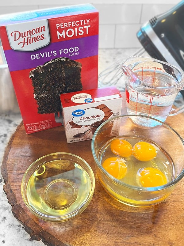 Photo of bowls of ingredients to make the chocolate cake batter for a coconut-filled Chocolate Bundt cake. On a wood riser is a box of Devil's Food chocolate cake mix (Duncan Hines) but you can use any box mix, a box of instant chocolate pudding (Great Value brand) again use any box mix, a measuring cup with one cup of water, a bowl containing eggs and egg whites and a small clear ingredient bowl with vegetable oil. In the background is a hint of a medium sized mixing bowl and a hand mixer.