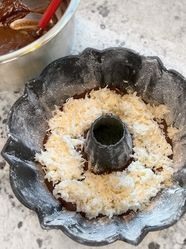 Process overhead photo of a Bundt pan filled with coconut filling mixture spread evenly over chocolate cake batter. In the background there is a hint of a mixing bowl with the remaining chocolate cake batter that will be next to spread on top.