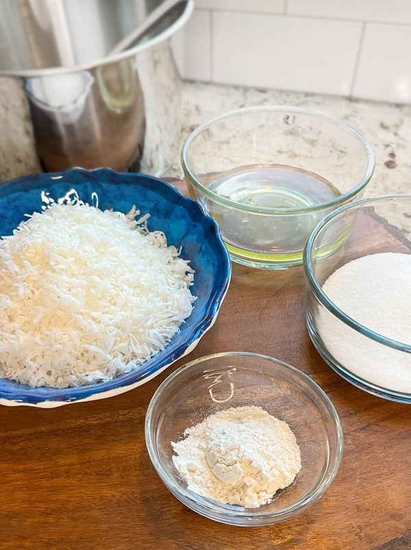 Photo of bowls of ingredients to make the coconut filling for a coconut-filled Chocolate Bundt cake. There is a medium sized stainless steel mixing bowl in the background. On a wood riser is a small ingredient bowl of flour. A larger blue bowl with flaked coconut, a bowl of egg whites and a bowl of sugar.