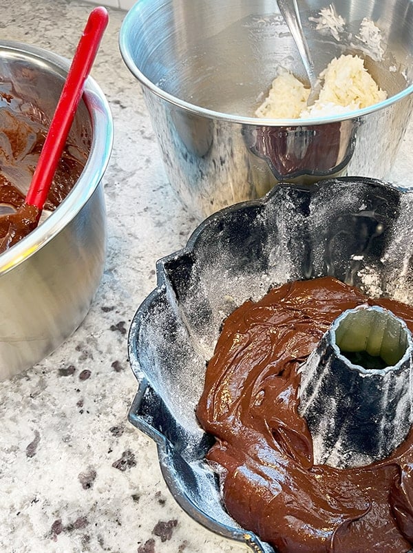 Process photo of the half of the cake batter that has been poured into a 10-inch Bundt pan. In the background are two stainless steel mixing bowls. One with the coconut filling mixture and the other hints at the remaining chocolate cake batter. This is the first step in layering ingredients in the Bundt pan to make a coconut filled chocolate Bundt cake.