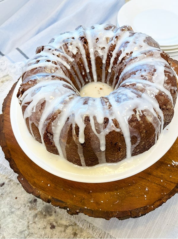 Chocolate Bundt cake just glazed. The glaze has been drizzled over the top and sides. The cake is on a white plate on a wood platter. In the background are white dessert plates.