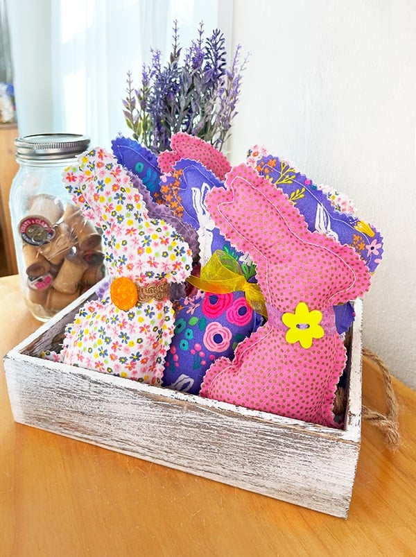Photo of a white wood crate with eight handmade stuffed bunnies in pinks polka dot, white floral and purple floral patterns for Easter. Some have bows around their neck and others have jute ribbon with a button embellishment. In the background is a ball jar with vintage wood spools and lilacs.