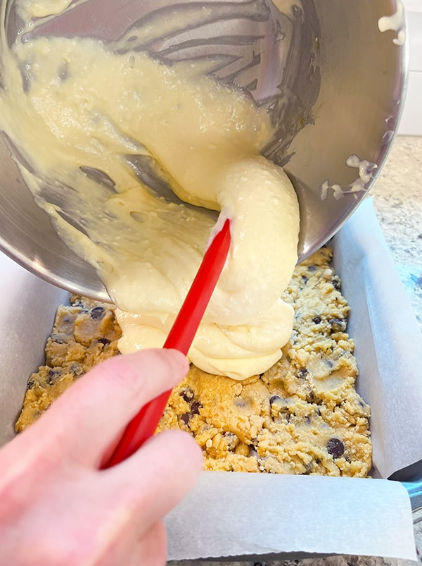 Cheesecake batter being poured from the mixing bowl over the chocolate chip cookie layer.