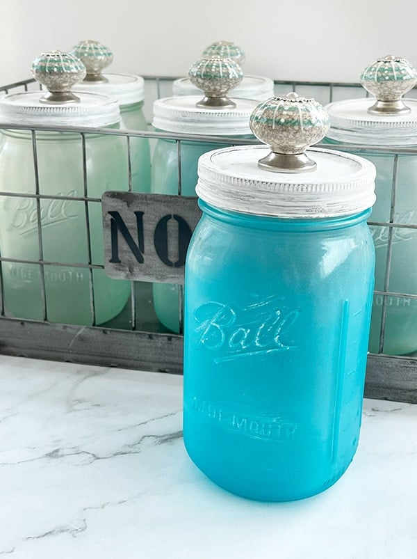 Vertical feature image of mason jars painted with sea glass spray paint in aqua and sea foam green. They are topped with a decorative knobs in gray and aqua that look like sea urchins. There are six jars. Five in a metal vintage crate and one in the foreground in aqua blue.