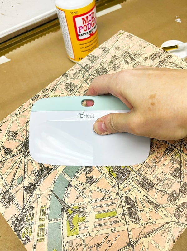 Using a squeegee to smooth out bubbles after using Mod Podge and a paper map panel to the top of a wood box.