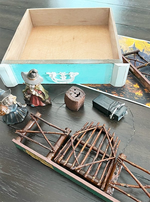 Photo of supplies used to make a Halloween miniature scene. These include an old small drawer, ceramic vintage witches, a metal mini jack-o-lantern, wood fence and a spooky scene printout to use as a background.