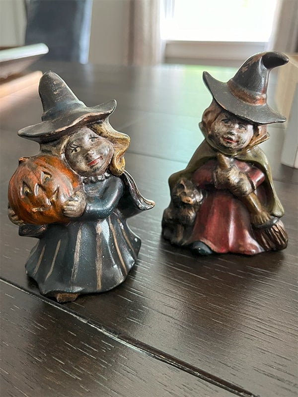 Photo of two ceramic vintage witches. Both are smiling and painted in muted dark blues, blacks and maroon. One holds a jack-o-lantern and the other has a broom with a black cat by her side. This miniature figures are about 3-inches tall.