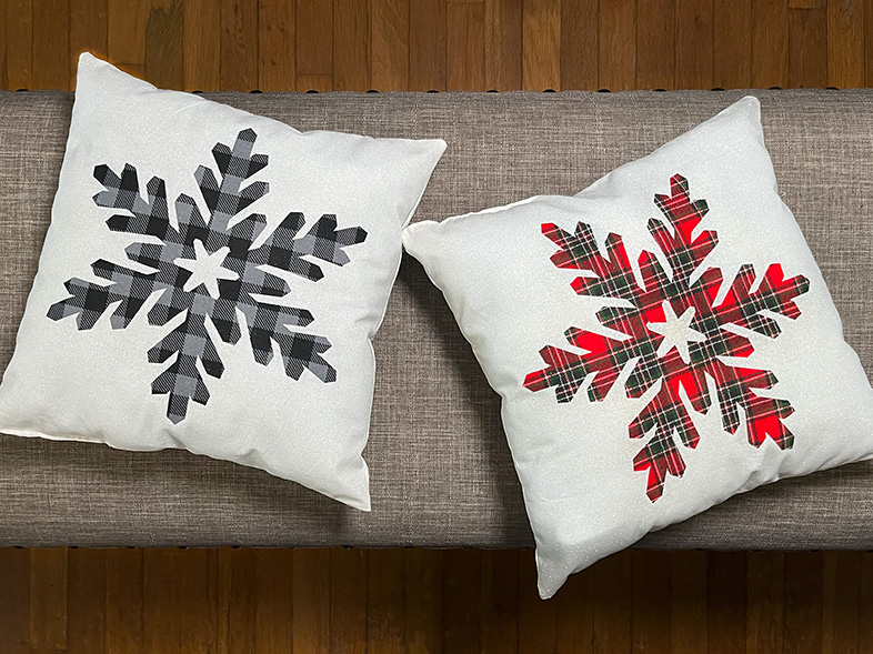 Photo of two white pillows with two different snowflake appliqued onto them. One has a red plaid snowflake and the other has a gray and black buffalo check snowflake.