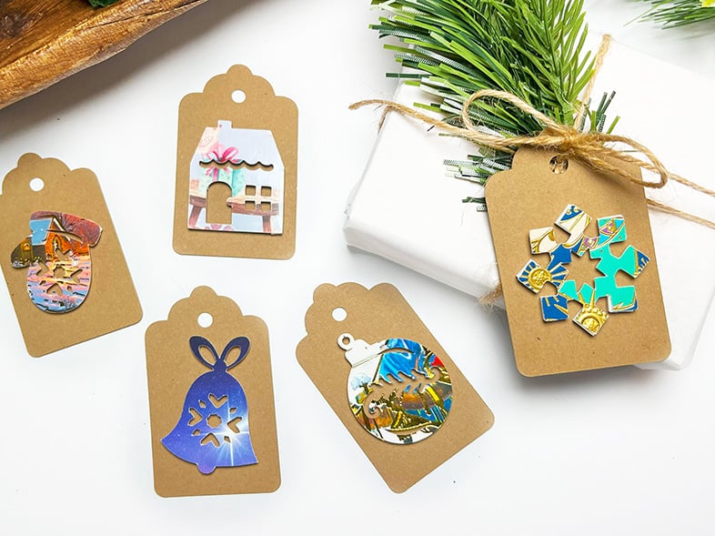 Horizontal photo with 4 finished recycled Christmas card gift tags on kraft card stock. One is on a white kraft paper wrapped gift tied with twine and a sprig of greenery. The other four tags are on the table next to the wrapped gift.