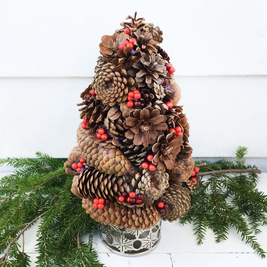 Square feature photo of a finished DIY Pinecone Christmas tree made with different varieties of pinecones. The tree is decorated with small red berries and below are spruce tree greenery. The tree sits in a silver snowflake cutout base.