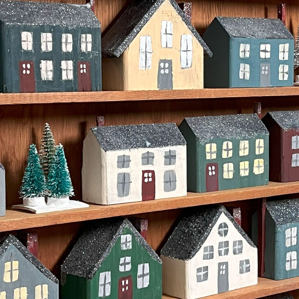Square feature photo of finished tiny wooden houses painted and on on a wood shelf. There are three shelves shown. The houses are painted dusty gues, greens, white and ivory. Each with dark gray roofs sprinkled with white glitter for a snowy effect. Beside some houses is a cluster of bottle brush trees.