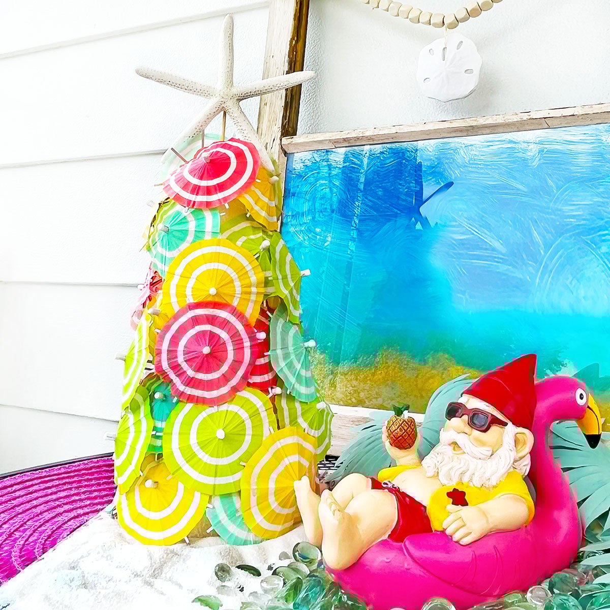 Square feature of a finished cocktail umbrella Christmas Tree with a starfish topper in a tablescape with a gnome holding a drink in a pineapple on a hot pink flamingo floatie. The colors of the umbrellas are bright pink, aqua, yellow and spring green.