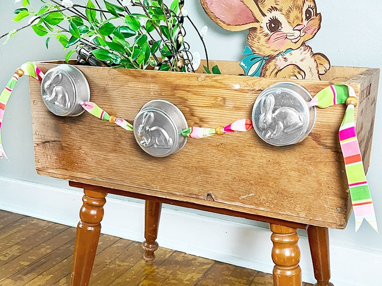 Bunny garland made from three round vintage bunny tart tins shown on a colorful spring ribbon. The ribbon has blocks of pink, green, spring green, fuchsia and orange. between the tins is a wood bead. This fun bunting is shown on the front of a vintage wood crate filled with spring greenery and a vintage bunny sign.