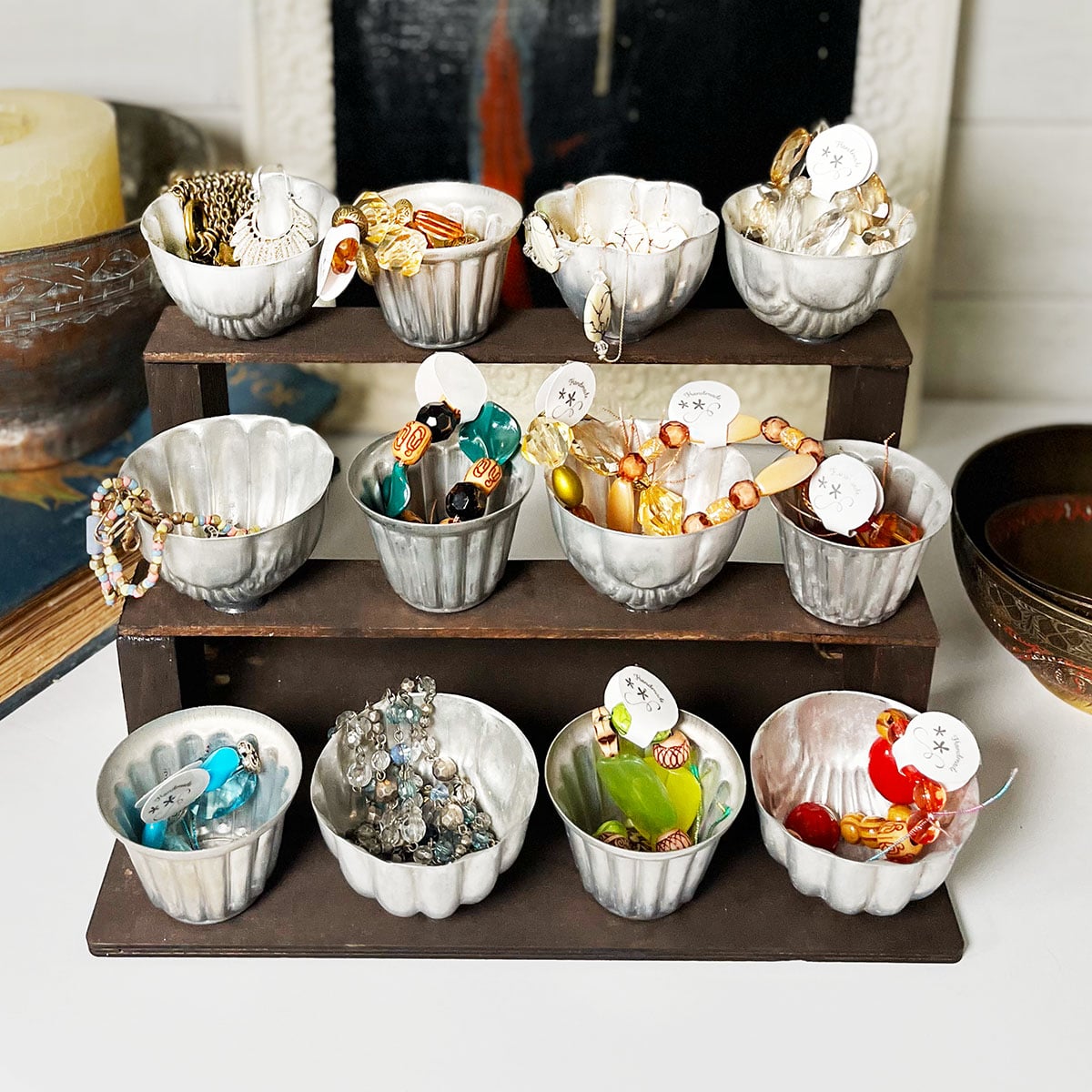 Square Feature image of a DIY three tiered organizer made with scrap wood and 12 vintage round and scalloped tart tins. There are four on each tier and they hold beads, small jewelry and bracelets with tags for sale in a vintage antique booth.