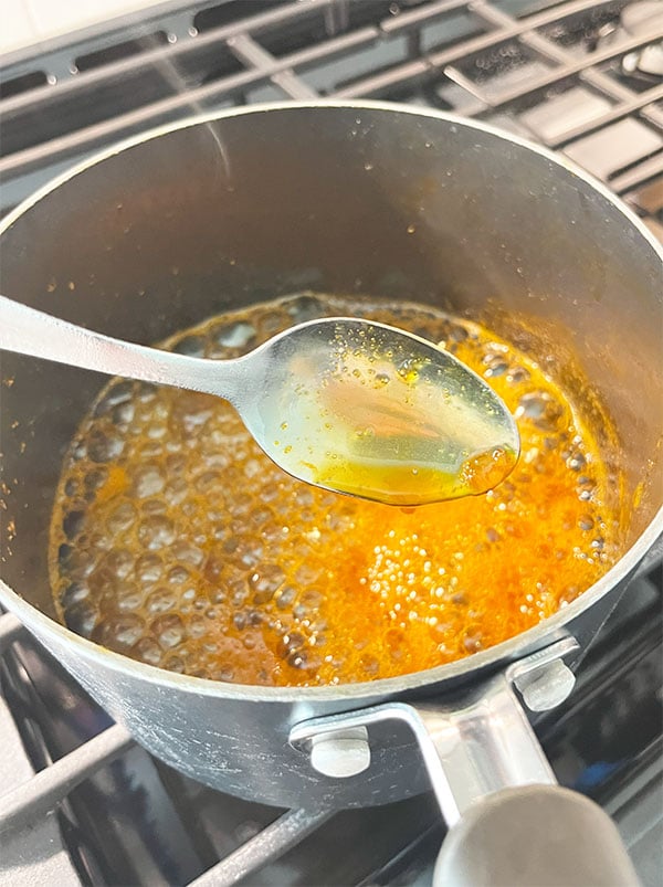 Closeup of a spoon dipped in a pan of orange glaze sauce thickening on the stove in a sauce pan. This shows the thickness needed. The sauce coats the spoon, slightly thickened.