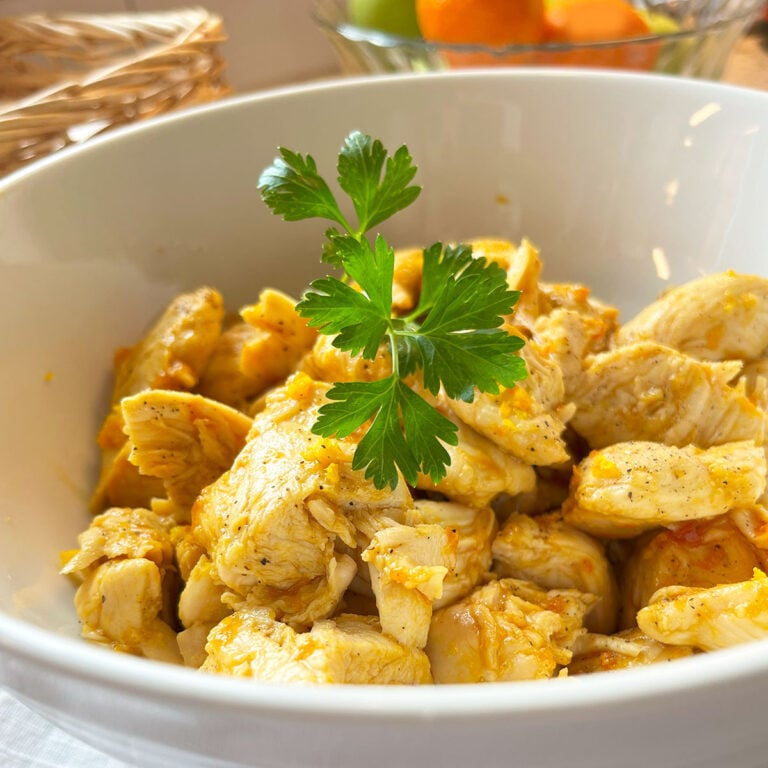 Square closeup feature image of a bowl of orange glazed chicken with a sprig of parsley on top in a white bowl. In the background is the hint of a bowl of oranges and a wicker basket.