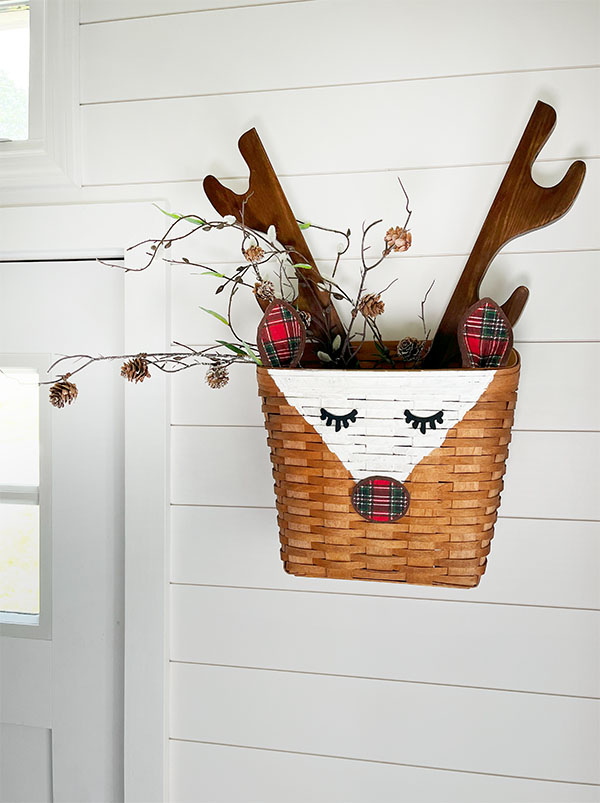 Square feature image of a square tapered wicker basket turned into a rustic reindeer basket with the addition of plaid red and green nose and ears and antlers made from an old vintage wall hanging gun rack. The basket is show here filled with a branch with small pine cones and branches of pussy willows hanging next to an entry way door on a white shiplap wall.
