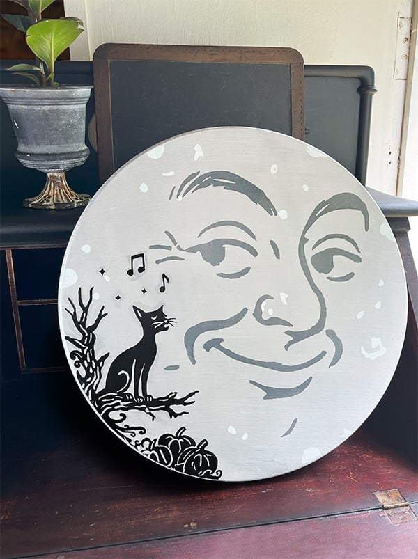 Round man in the moon sign - Vintage style stenciled sign with a black cat singing, sitting on a knotty branch with pumkins below. The moon man face take up more than 3/4 of the round sign with the cat in the left corner. The colors are light gray for the moon, darker gray for the man face on the moon and black silhouette style for the cat scene. This fall sign was stenciled using cut stencil vinyl cut on a Cricut Maker 3.