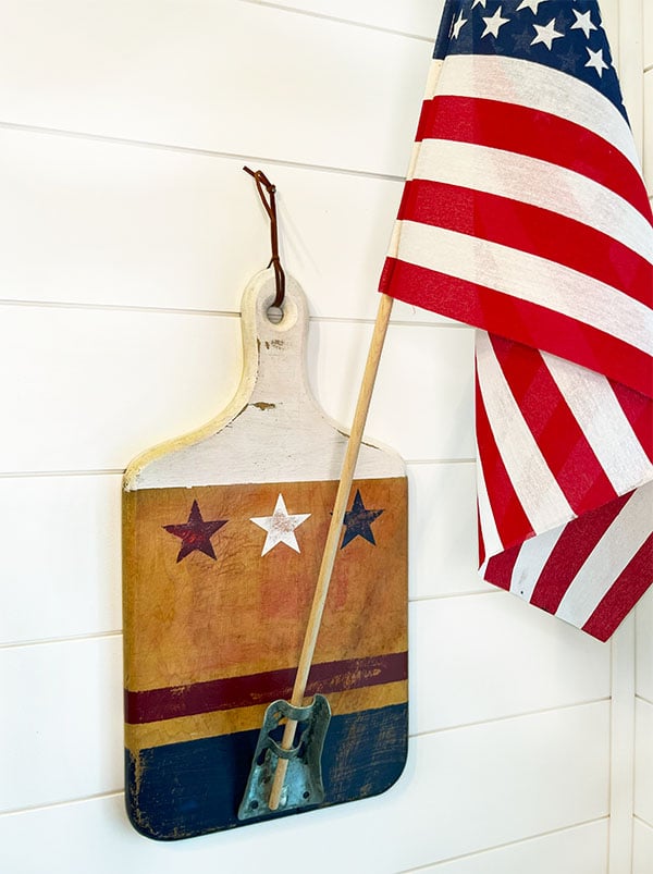 Wood handle cutting board shown hanging by a leather cord on a white wood wall. The cutting board has been painted white at the top with a red, white and blue star below. At the bottom is a red stripe across horizontally and below that the bottom portion is painted blue. At the bottom center is a metal flag holder with a tall United States flag in the holder.