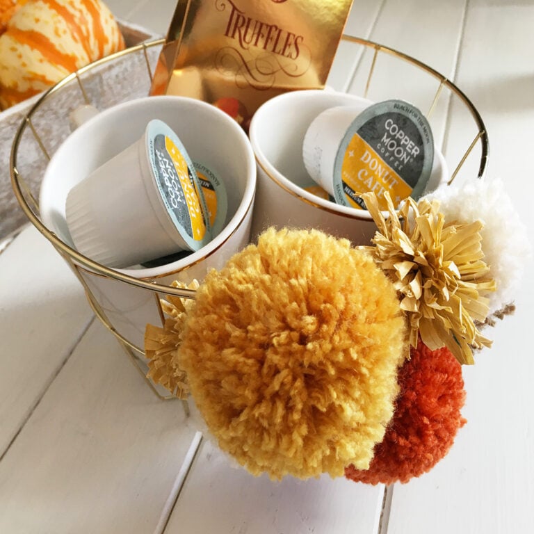 Easy Fall Wire Basket Makeover with Pom Poms - a gold Dollar Tree round wire basket with gold, cream, rust and raffia textured pom poms on the front shown on white background filled with two white coffee mugs, specialty coffee pods and a foil box of truffles to give as a gift basket.