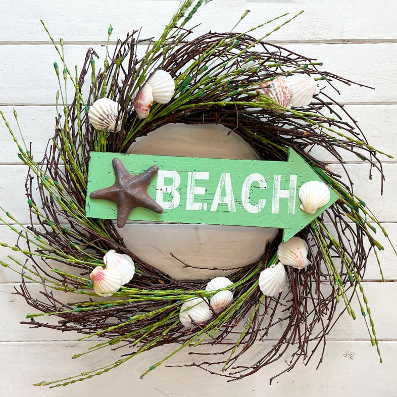 Beachy wreath made from a thrifted twig wreath with green grasses combined with the twigs. In the center is a wood arrow shaped sign in a light green that has a metal shell and says 'BEACH'. The wreath and beach sign came from the thrift store and I just hot glued shells randomly to the twig wreath.
