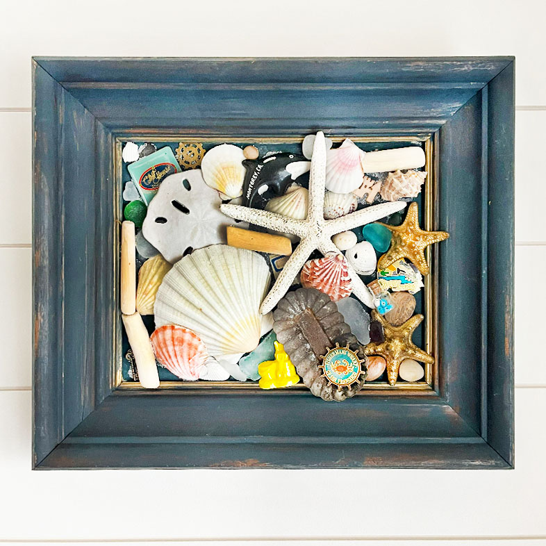 Shown is a dark blue wood frame with beach items and tiny trickets, there are seashells, starfish, sea glass, magnets, beach finds like smooth pebbles and a rusty tin. All arrange in a collage with a sprinkle sand glued in the some of the spaces.