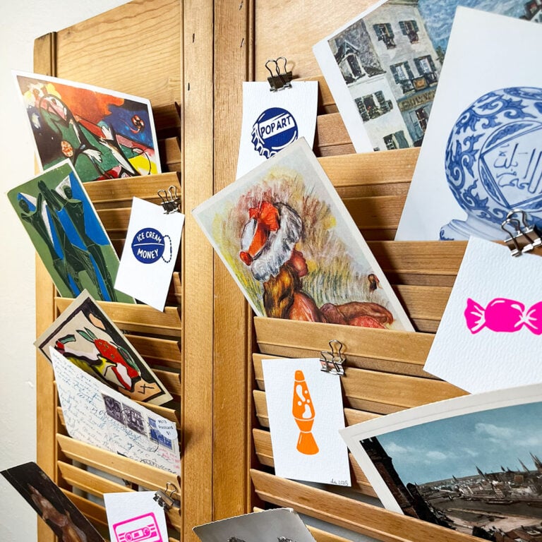 Closeup at a 3/4 angle of a DIY card holder made from old shutters. Here I filled it with vintage postcards and colorful, handmade 2x3 screen prints of vintage items like a coin purse that says ice cream money, a lava lamp and wrapped candy.